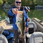 Angler proudly holding a large bass caught on Lac Heney while standing inside a fishing boat on a calm summer morning.